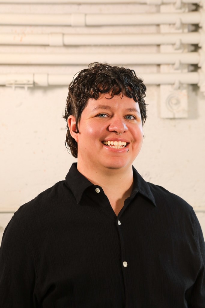 Portrait of Ray Martinez who is wearing a black button up shirt and smiling while standing in front of a white wall. 